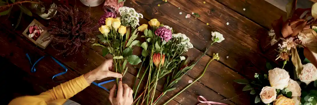 person in yellow shirt arranging flowers including yellow tulips and white roses on a brown wooden table